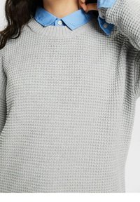 Lichtgrijze gebreide trui met een ronde hals, gedragen over een blauwe blouse met kraag, met knoopdetails op de kraag.