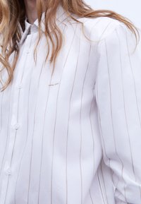 Camicia bianca con bottoni, decorata con sottili righe beige, realizzata in tessuto morbido, dotata di colletto classico e un solo dettaglio cucito sul petto.