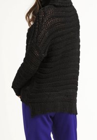 Maglione nero lavorato a maglia con un motivo testurizzato, caratterizzato da una vestibilità ampia e un collo alto. L'orlo è più lungo sul retro, in perfetta armonia con i pantaloni blu.