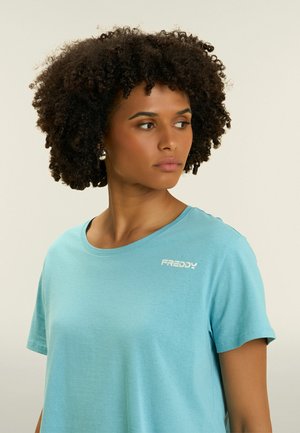 Femme aux cheveux bouclés portant un t-shirt bleu clair de la marque Freddy, regardant sur le côté sur un fond uni.