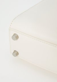 Sac en cuir synthétique couleur crème avec une texture lisse, des coins arrondis, des coutures contrastantes et deux pieds en métal au fond.
