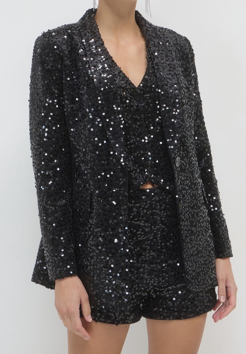 Blazer noir à sequins et short à sequins assorti portés par une personne sur un fond clair uni.