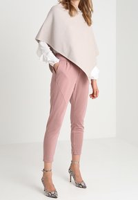 Poncho rose pâle en tissu texturé sur une chemise blanche, associé à un pantalon rose fuselé et des talons en serpent argenté avec des brides de cheville.