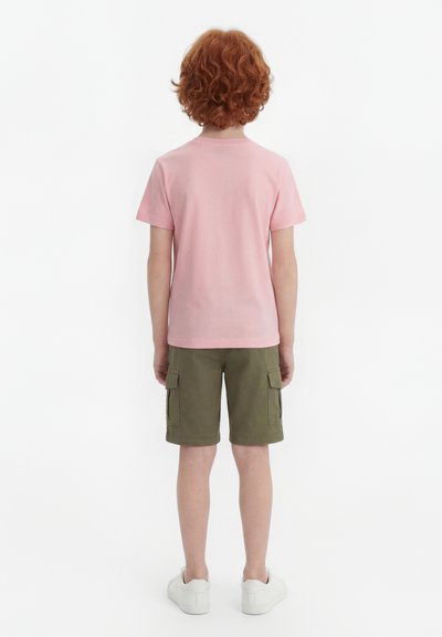 Παιδί με σγουρά κόκκινα μαλλιά που φοράει ροζ μπλουζάκι, χακί cargo σορτς και λευκά αθλητικά παπούτσια, στέκεται με την πλάτη προς τα εμπρός σε λευκό φόντο.