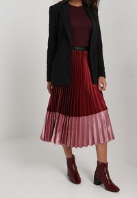 Zwarte blazer over een kastanjebruine top, gecombineerd met een geplooide rok met een verloop van donkerrood naar zachtroze. Glanzende rode enkellaarzen.