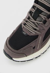 Kratak prikaz smeđih i crnih tenisica Armani Exchange sa teksturiranim mrežastim materijalom, detaljima od antilopa, označenim etiketa i bež potplatom.