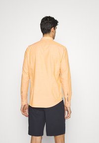Hombre con cabello corto rizado oscuro, vistiendo una camisa de manga larga color melocotón y pantalones cortos negros, de pie de espaldas contra un fondo blanco.
