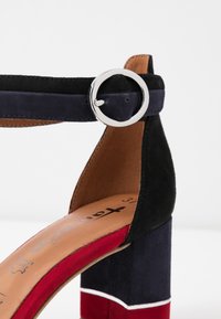 Czarno-niebieskie zamszowe sandały na słupku z okrągłą srebrną klamrą, czerwonymi i białymi akcentami na obcasie oraz brązową skórzaną wkładką.