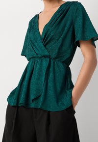 Blusa avvolgente turchese con maniche a sbuffo corte, tessuto testurizzato e vita cinchata, abbinata a pantaloni neri. Design semplice e silhouette fluida.