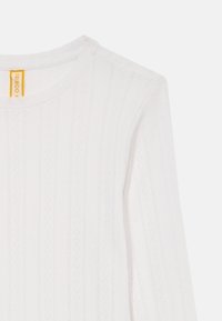 Biela dlhá mikina s dlhými rukávmi, ktorá má textúrovaný vzor vertikálnych pruhov a diamantov, okrúhly výstrih a žltú značkovú etiketu vo vnútri.