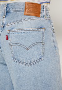 Světlé modré džíny z denimu s hnědou koženou nášivkou, které mají charakteristický design zadní kapsy a kontrastní oranžové prošívání.