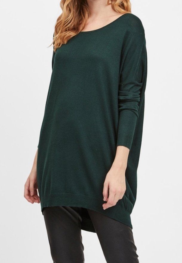 Femme portant un pull ample vert foncé à manches longues et un pantalon noir, debout devant un fond uni.