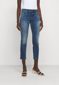 Jeans de mezclilla en azul medio, de tiro medio con pierna ajustada, ligera decoloración y dobladillo deshilachado. Combinados con tacones blancos con tiras.