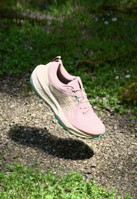 Gaiši rozā sporta apavi ar iemecu, sietiņu augšdaļu, dzelteniem un ziliem akcentiem, kā arī teksturētu dizainu, kas karājas virs grants virsmas.