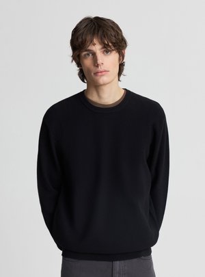 Giovane uomo con capelli castani di media lunghezza che indossa un maglione nero girocollo e pantaloni grigi, in piedi davanti a uno sfondo chiaro e uniforme.