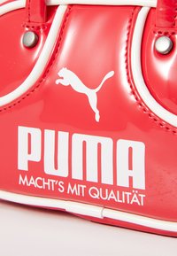 Röd glansig handväska med vita accenter. Har djärv "PUMA"-logotyp och texten "MACHT'S MIT QUALITÄT" i stora vita bokstäver.
