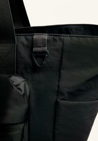 Fekete, texturált felületű tote táska oldalzsebbel, állítható pánttal és fémszeraffal a tárgyak rögzítéséhez. Könnyű anyagból készült.