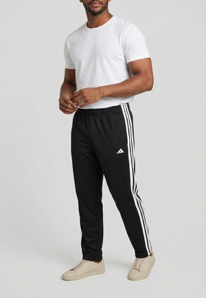 Hombre vistiendo una camiseta blanca, pantalones deportivos negros Adidas con franjas blancas y zapatillas beige, de pie frente a un fondo liso.