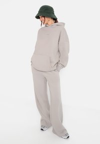 Felpa grigia chiara over-size con tasca a marsupio, abbinata a pantaloni della tuta a gamba larga dello stesso colore. Cappello a secchiello verde testurizzato e sneakers.