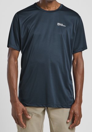 Uomo che indossa una t-shirt a maniche corte Jack Wolfskin blu scuro e pantaloni beige, in piedi con le braccia rilassate ai lati.