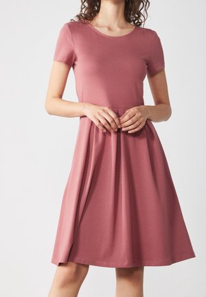 Kvinde iført en kortærmet, knælængde kjole i støvet rosa med taljeret pasform og udvidet skørt, stående mod en ensfarvet lys baggrund.