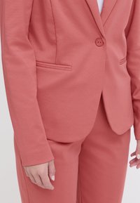 Koraalblazer met een enkele knoop, twee broekzakken en een fitted design. Gemaakt van textuurstof, gecombineerd met bijpassende broek.