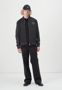 Black bomber jacket with a textured finish, silver zipper, and logo patch, paired with a black shirt, trousers, and cap. Smooth fabric.