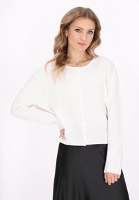 Bílý pletený kardigan s kulatým výstřihem, zapínáním na knoflíky a dlouhými rukávy. Hladká textura, zkrácená délka, kombinován s černou sukní.