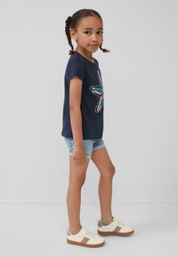 Marineblauw t-shirt met een kleurrijke sterafbeelding, lichte denim shorts en witte sneakers met bruine accenten en een grijze tong.