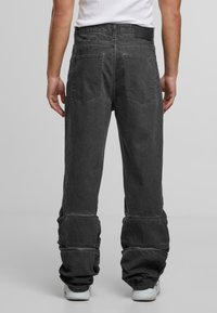 Osoba v černých volných džínách s viditelným šitím a velkými zadními kapsami, v kombinaci s bílými teniskami a bílým tílkem.