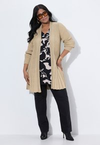 Mujer con un cárdigan beige de canalé, una blusa floral en blanco y negro, pantalones negros, botas de tacón negras, gafas de sol grandes y pendientes de aro dorados.