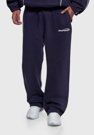 Osoba w luźnych granatowych dresach z białym logo „DROPSIZE” i białych sneakersach, stojąca na jednolitym tle.