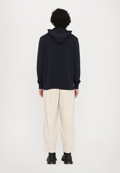 Sudadera navy con cordón, combinada con pantalones de pierna ancha color beige claro y zapatos negros, vista desde atrás sobre un fondo plano.