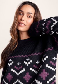 Young woman with long brown hair wearing a black sweater with white and purple geometric patterns, posing with one arm raised behind her head.