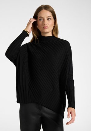 Vrouw met steil haar die een geribde zwarte trui en zwarte broek draagt, die met één hand haar haar aanraakt tegen een effen lichte achtergrond.