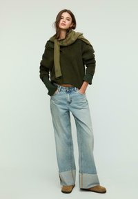 Maglione verde a maglia con maniche oversize, abbinato a jeans in denim blu chiaro a gamba larga con orlo grezzo. Stivali marroni alla caviglia completano l'outfit.