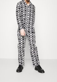 Person i matchande svart- och vitfärgad långärmad skjorta och byxor med svarta kraftiga snörskor mot en enkel bakgrund.