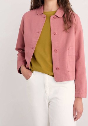 Person trägt eine pinke, geknöpfte Jacke über einem senfgelben Hemd, mit einer Hand in der weißen Hosentasche, steht vor einem hellen Hintergrund.