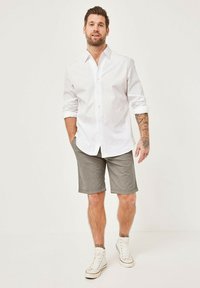 Weißes, knopfbares Hemd, graue gemusterte Shorts und weiße High-Top-Sneaker. Legerer Stil mit hochgekrempelten Ärmeln und sichtbaren Bein-Tattoos.