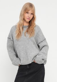 Maglione grigio a coste con maniche ampie e cuciture decorative lungo le spalle, abbinato a una gonna in denim scuro.