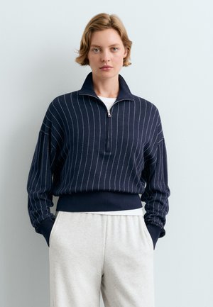 Frau mit kurzen blonden Haaren, die einen marineblauen Reißverschluss-Pullover mit weißen Streifen und hellgraue Hose trägt, steht vor einem einfarbigen hellen Hintergrund.