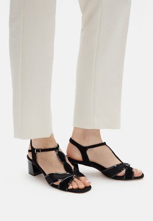 Czarne sandały na obcasie wykonane z lakierowanej skóry, z paskiem w kształcie litery T, niskim obcasem typu bloczek oraz skrzyżowanymi paskami na palcach.