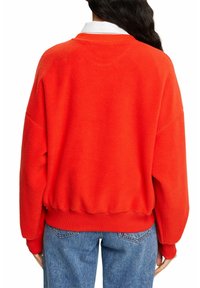 Pull-over en polaire rouge avec une coupe décontractée, épaules tombantes, poignets et ourlet côtelés. Porté sur une chemise blanche à col, associé à un jean bleu.