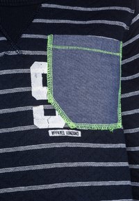 Námořnicko-modré a bílé pruhované tričko s krátkým rukávem, se strukturovanou kapsou, zeleným prošíváním a bílým číslem "9" tištěným na přední straně.