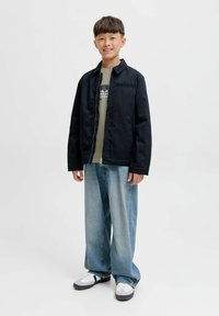 Dečko sa smiješkom nosi crnu jaknu, majicu s printom, široke svijetloplave traperice i bijele tenisice, stoji ispred jednolične pozadine.