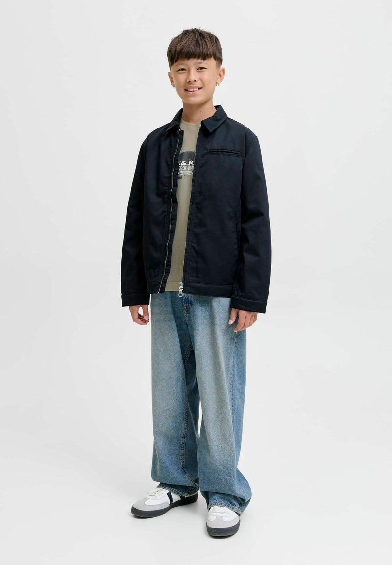 Dečko sa smiješkom nosi crnu jaknu, majicu s printom, široke svijetloplave traperice i bijele tenisice, stoji ispred jednolične pozadine.