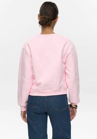 Rozā kokvilnas sporta krekls ar apaļu kaklu, nokaltām plecu šuvēm un ribinātām piedurknēm. Apvienots ar tumši zilām svītrainām džinsu biksēm.