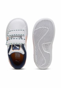 Baskets blanches avec deux bandes Velcro, doublure bleu marine et accents orange. Semelle blanche texturée avec un motif en zigzag. Logo Puma visible.
