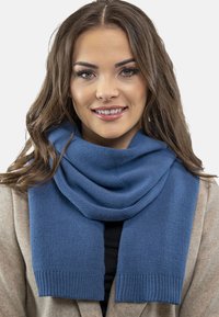 Kék kötött sál, puha tapintással, bordázott széllel és laza eséssel, bézs kabát felett viselve.