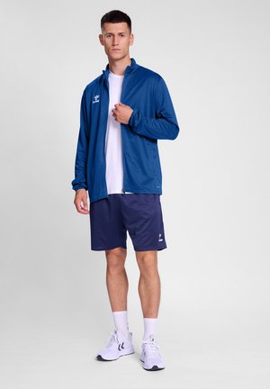 Blauwe sportjacker, witte t-shirt, marineblauwe shorts en witte sneakers. De jacker heeft een hoge kraag en ritsdetails, ontworpen voor actieve kleding.